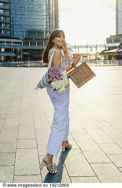 Happy woman walking with bouquet of flowers and basket on footpath