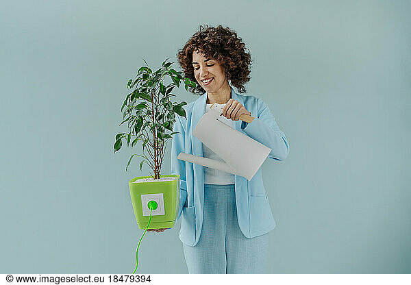 Happy woman watering potted plant connected with electric plug against blue background
