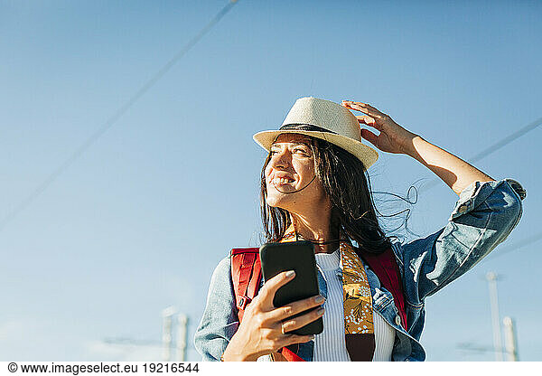 Happy woman wearing hat and standing with smart phone under sky