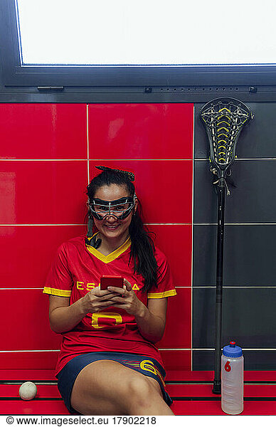 Happy woman wearing protective eyewear sitting with mobile phone in locker room