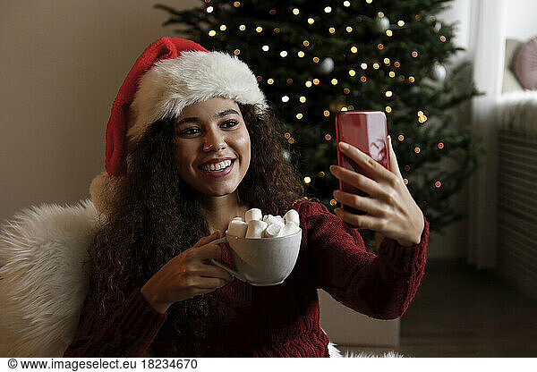 Happy woman wearing Santa hat and taking selfie with cup of marshmallow cocoa