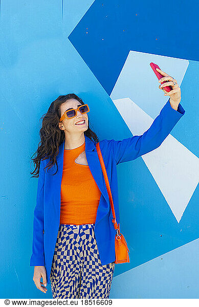 Happy woman wearing sunglasses taking selfie in front of blue wall