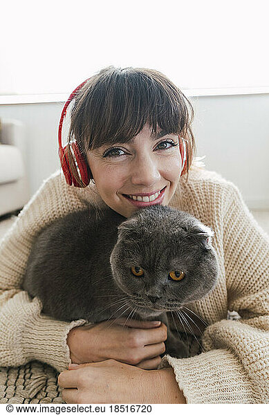 Happy woman wearing wireless headphones embracing cat at home