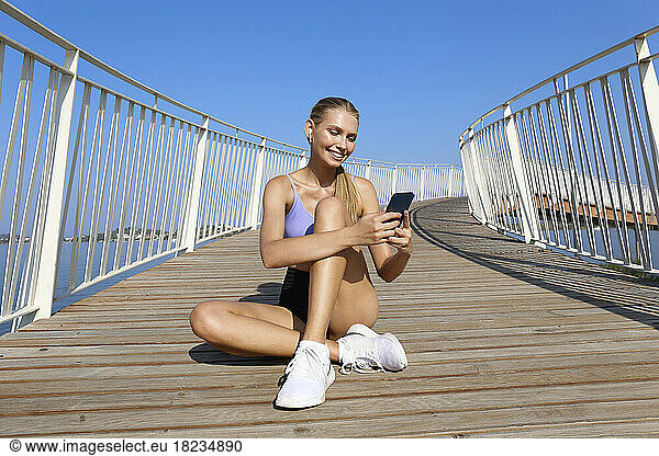 Happy woman wearing wireless in-ear headphones looking at mobile phone on elevated walkway