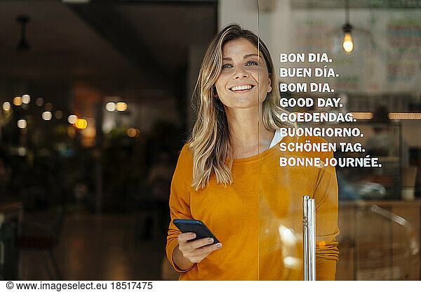 Happy woman with blond hair holding smart phone standing at door