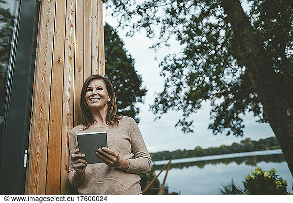 Happy woman with brown hair standing with tablet PC by wooden wall at backyard