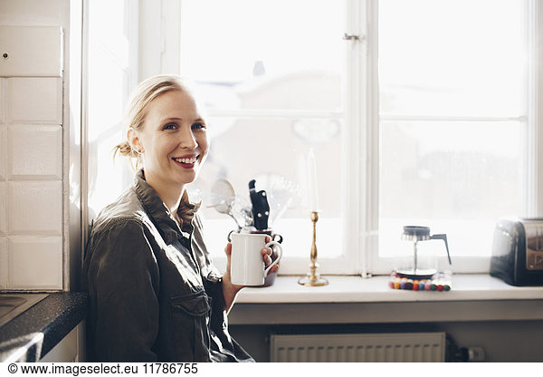Happy woman with coffee cup against window in kitchen at home