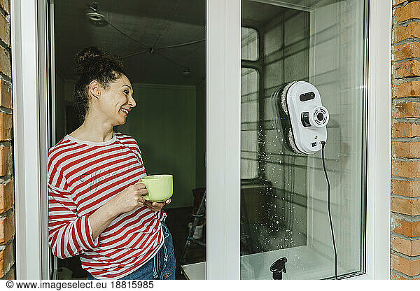 Happy woman with coffee cup cleaning window through robot washer