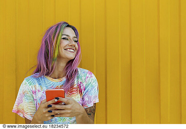 Happy woman with colorful hair holding mobile phone in front of yellow wall