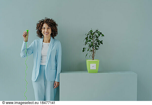 Happy woman with electric plug standing by potted plant in front of blue wall