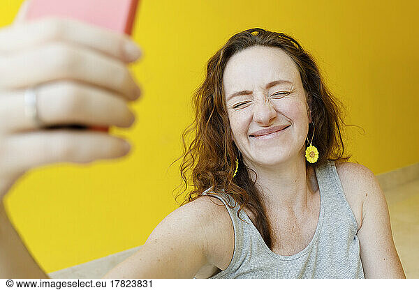 Happy woman with eyes closed taking selfie through mobile phone in front of yellow wall
