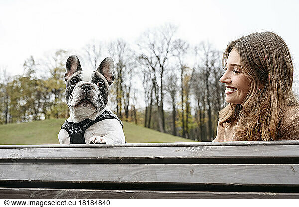 Happy woman with French bulldog sitting on bench at park