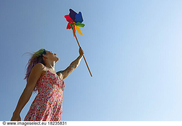 Happy woman with hand raised holding colorful pinwheel toy gesturing under blue sky