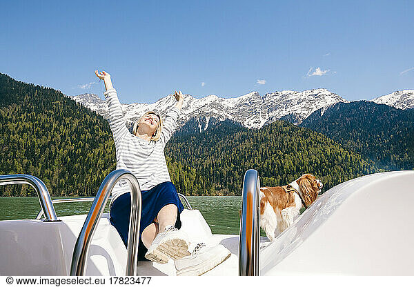 Happy woman with hands raised sitting by dog on boat