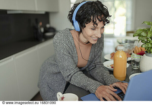Happy woman with headphones working from home at laptop in kitchen
