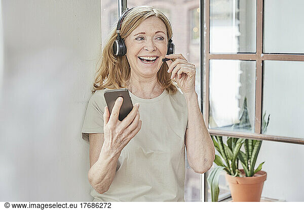 Happy woman with headset and smart phone in front of window at home