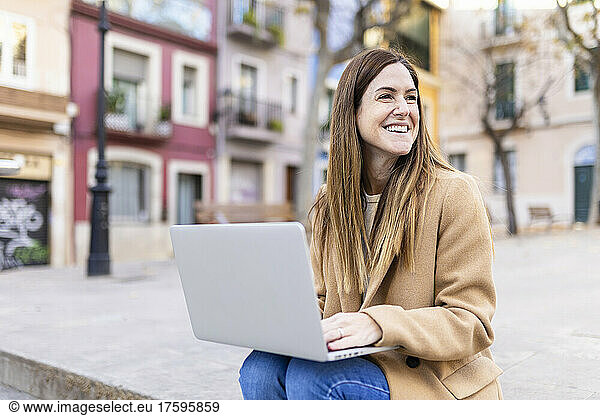 Happy woman with laptop sitting on footpath in city