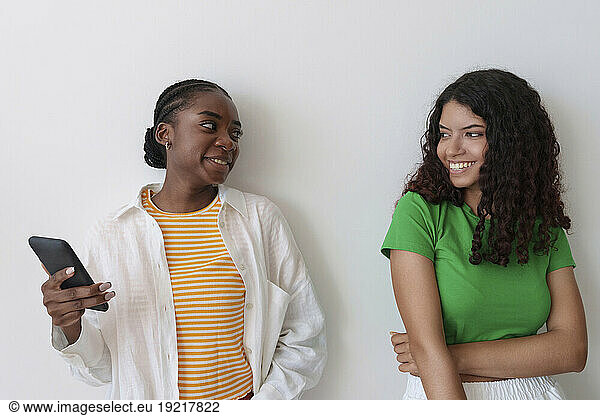Happy woman with smart phone looking at friend in front of wall