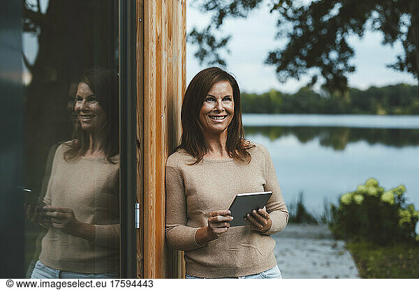 Happy woman with tablet PC standing by glass wall at backyard