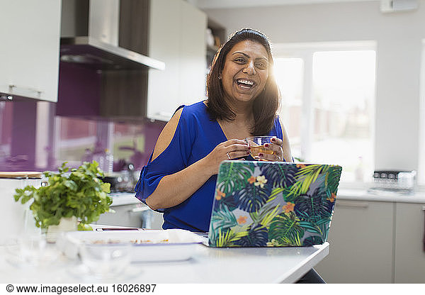 Happy woman with tea laughing at laptop in kitchen