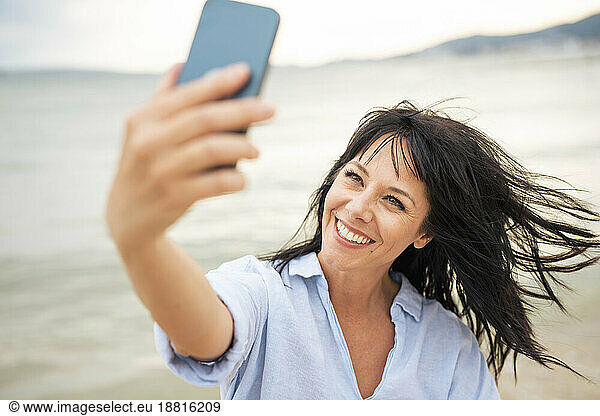 Happy woman with tousled hair taking selfie through smart phone at beach