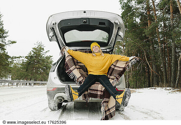 Happy woman with warm clothing sitting in car trunk at winter