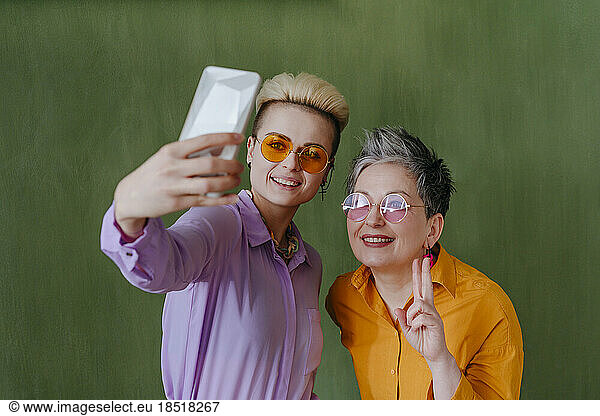 Happy women taking selfie through smart phone in front of green wall