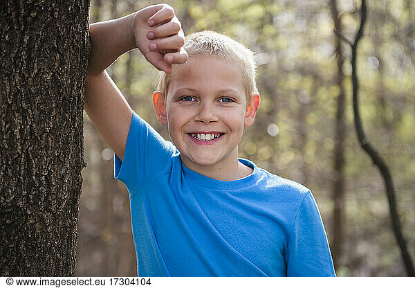 Happy young blond boy leaning against a tree in the woods.