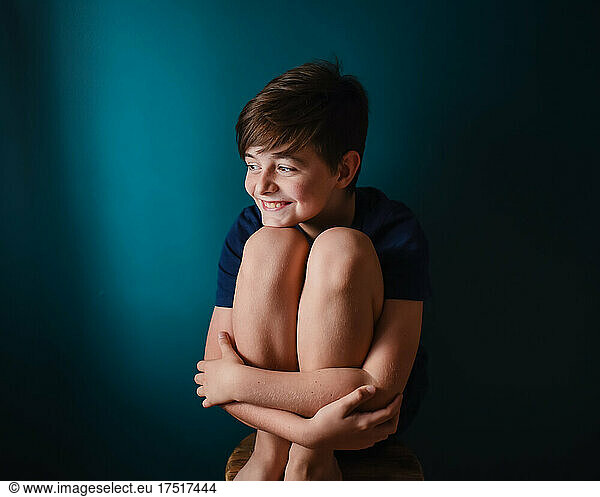 Happy young boy sitting on a stool against a dark blue wall.