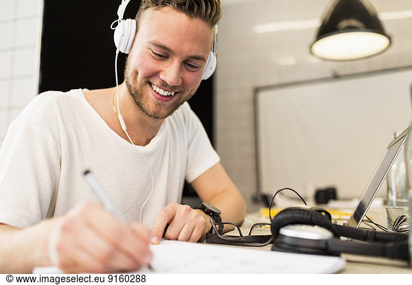Happy young businessman wearing headphones while writing in book at creative office