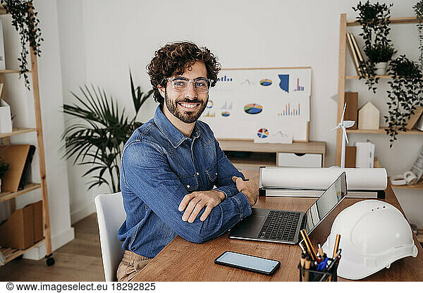 Happy young engineer wearing eyeglasses sitting with laptop at desk in office