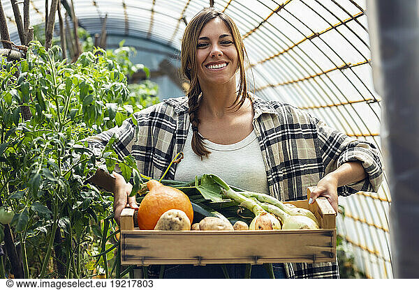 Happy young farm worker with freshly harvested vegetables in wooden crate at greenhouse