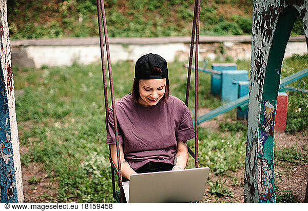 Happy young female student using laptop sitting on a swing