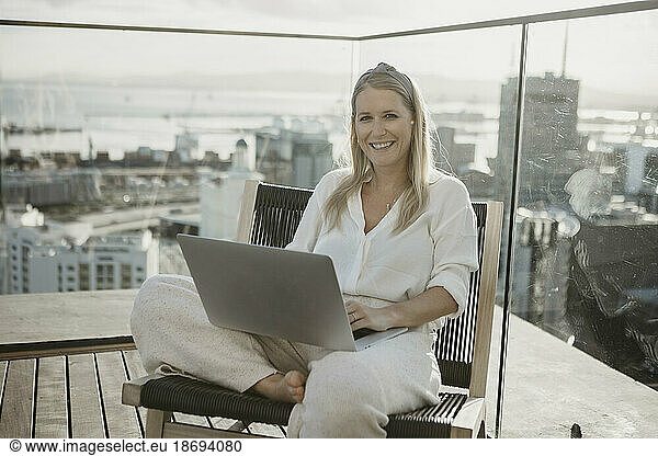 Happy young freelancer with laptop sitting on chair on rooftop