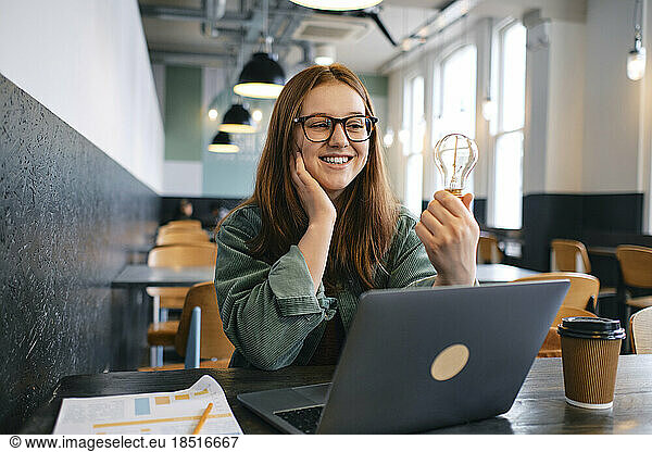 Happy young freelancer with light bulb sitting in cafe