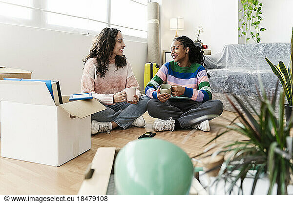 Happy young friends sitting with coffee cup on floor at home