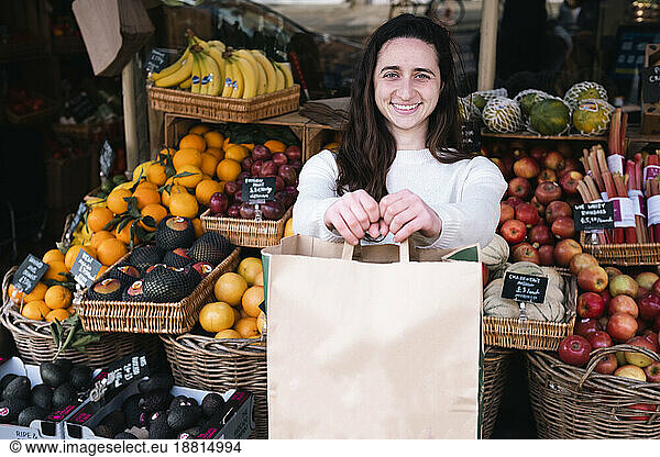 Happy young grocer standing with paper bag at vegetable shop