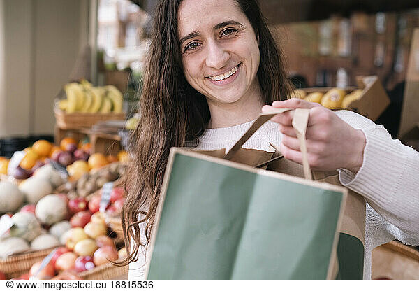 Happy young grocer standing with paper bag at vegetable stall