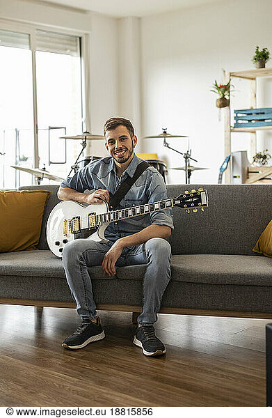 Happy young guitarist sitting with guitar on sofa at home