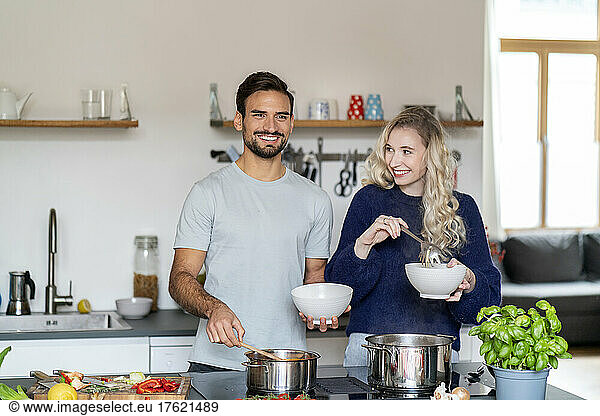 Happy young man and woman holding bowls standing in kitchen at home