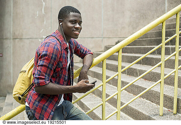 Happy young man listening to music holding smart phone leaning on railing