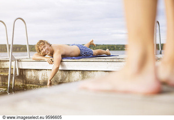 Happy young man looking at friend while lying on boardwalk against sky