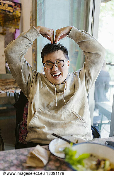 Happy young man making heart shape with hands while sitting at restaurant