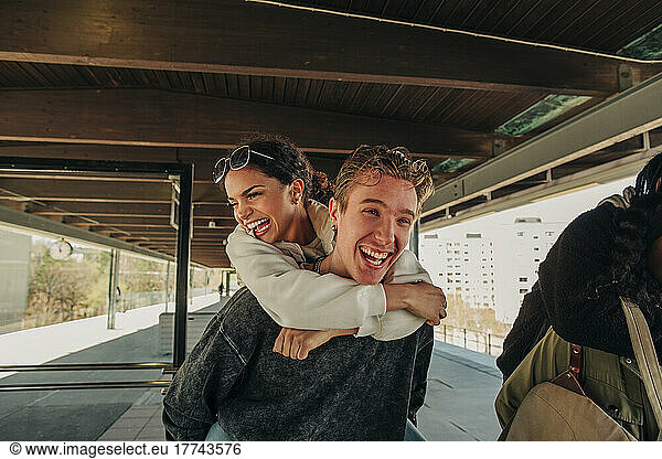 Happy young man piggybacking female friend at railroad station platform