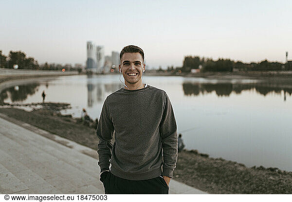 Happy young man standing with hands in pockets by lake at sunset