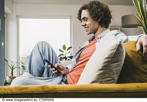 Happy young man surfing net through mobile phone sitting on sofa at home
