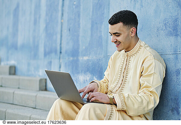 Happy young man using laptop sitting on steps by blue wall