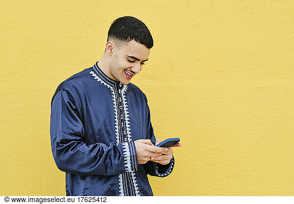 Happy young man using mobile phone standing in front of yellow wall