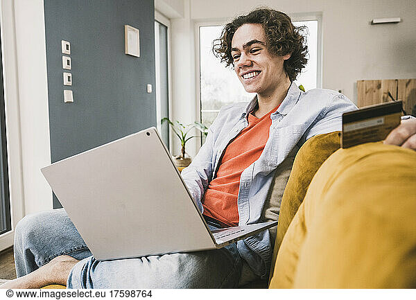 Happy young man with credit card doing online shopping through laptop at home