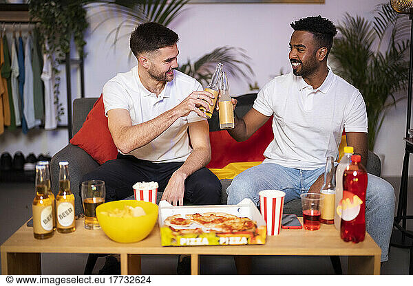 Happy young men toasting beer bottles sitting on sofa in living room at home
